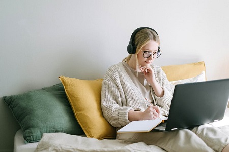 Chica joven estudiando en el ordenador desde el sofa y tomando notas